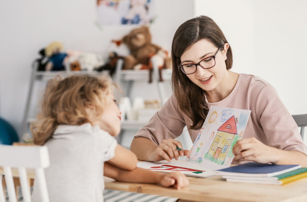 Psicóloga infantil ajudando uma criança a compreender atividades educativas com um sorriso, sentada numa sala de aula.